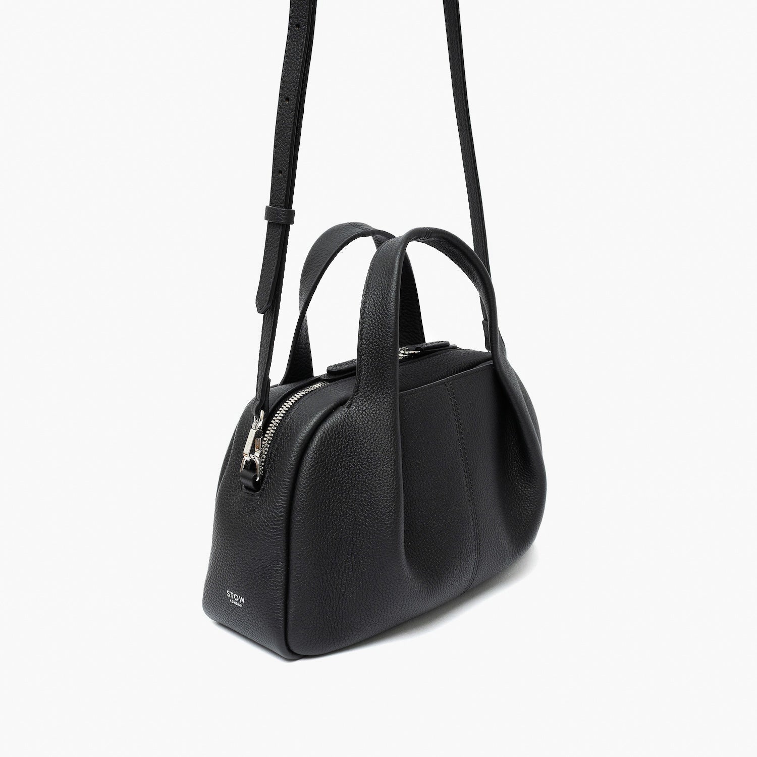 Off-axis view of STOW London's Mini Bowling bag in Black pebbled leather featuring a pleated front panel, detachable crossbody strap and silver stainless steel hardware on a light grey background