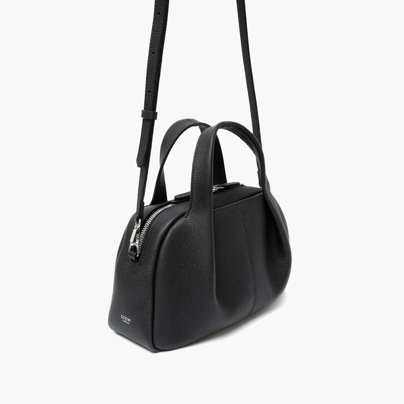 Off-axis view of STOW London's Mini Bowling bag in Black pebbled leather featuring a pleated front panel, detachable crossbody strap and silver stainless steel hardware on a light grey background
