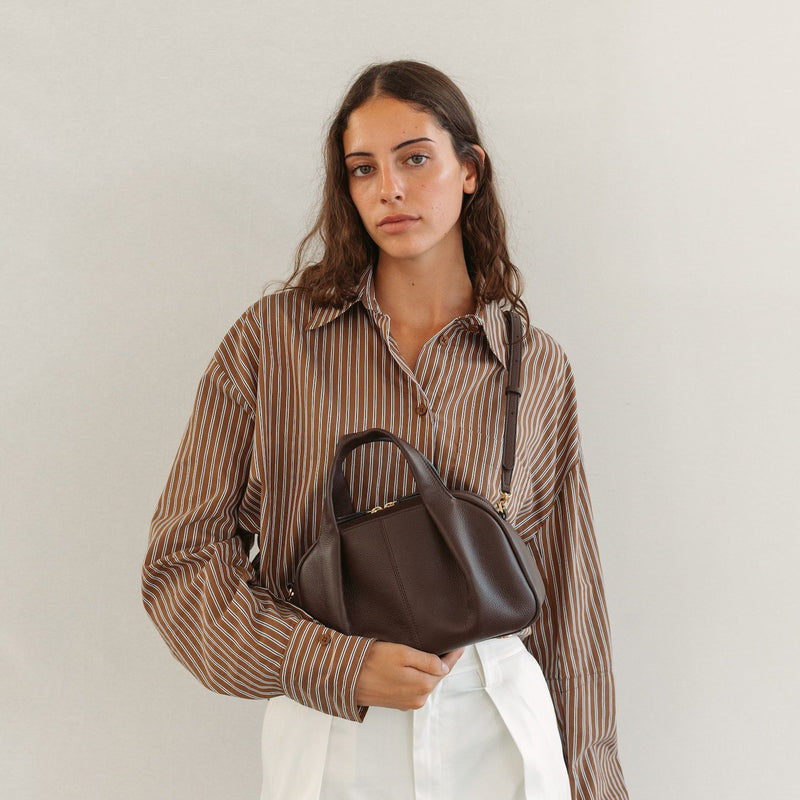Model holding STOW London's Mini Bowling in Espresso pebbled leather featuring a pleated front panel and gold-coated stainless steel hardware 