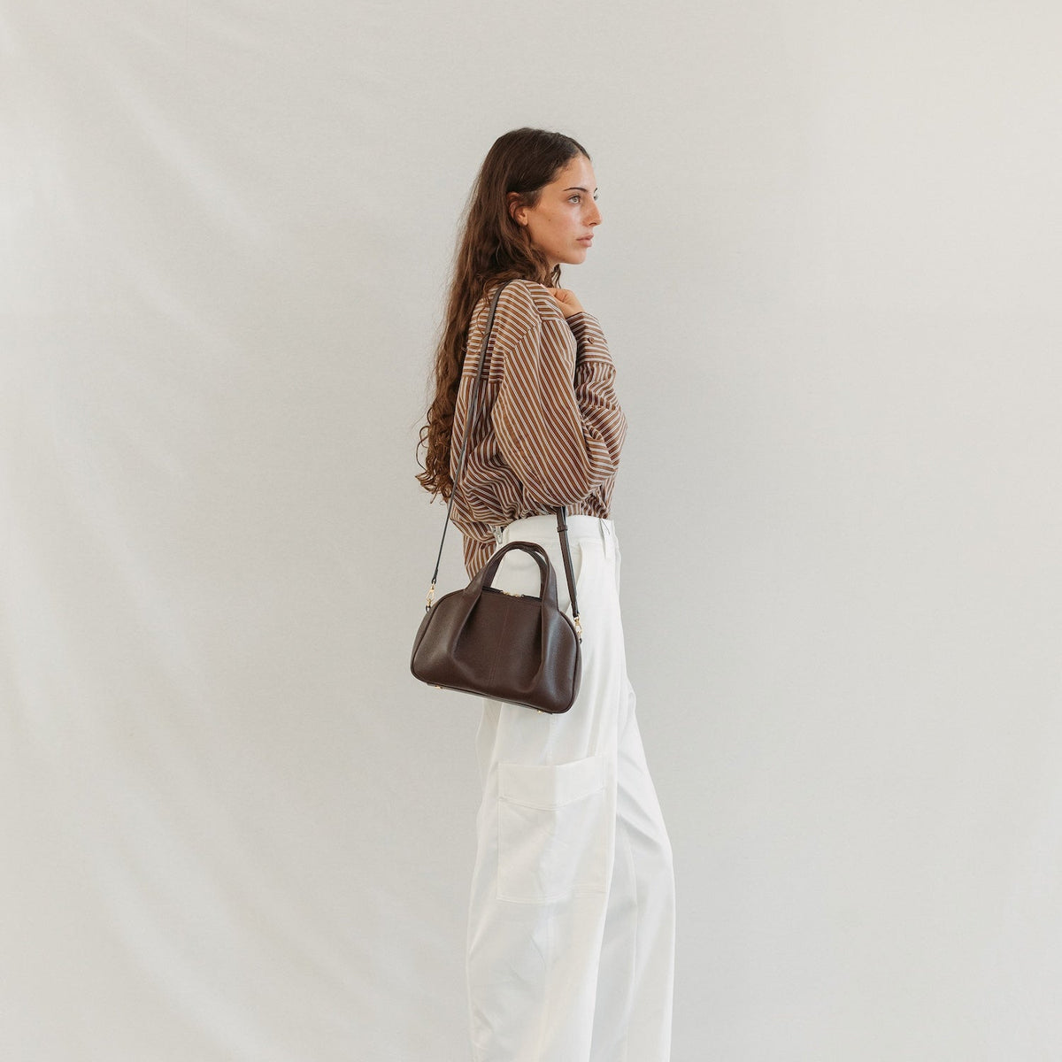 Model wearing STOW London's Mini Bowling in Espresso pebbled leather crossbody featuring a pleated front panel and gold-coated stainless steel hardware 