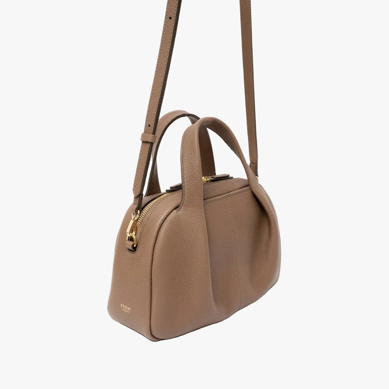 Off-axis view of STOW London's Mini Bowling in Mocha pebbled leather with its crossbody strap attached featuring a pleated front panel and gold-coated stainless steel hardware on a light grey background 