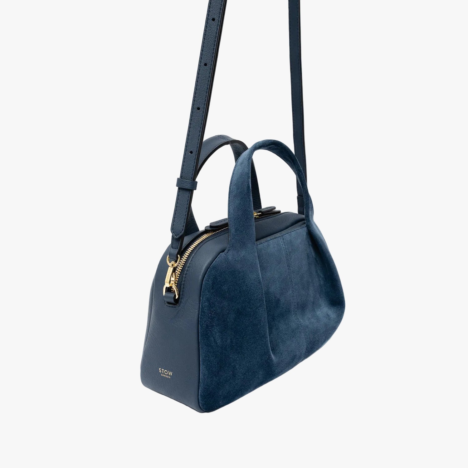 Off-axis view of STOW London's Mini Bowling bag in Navy suede and smooth leather featuring its detachable crossbody strap, a pleated front panel and gold-coated stainless steel hardware on a light grey background