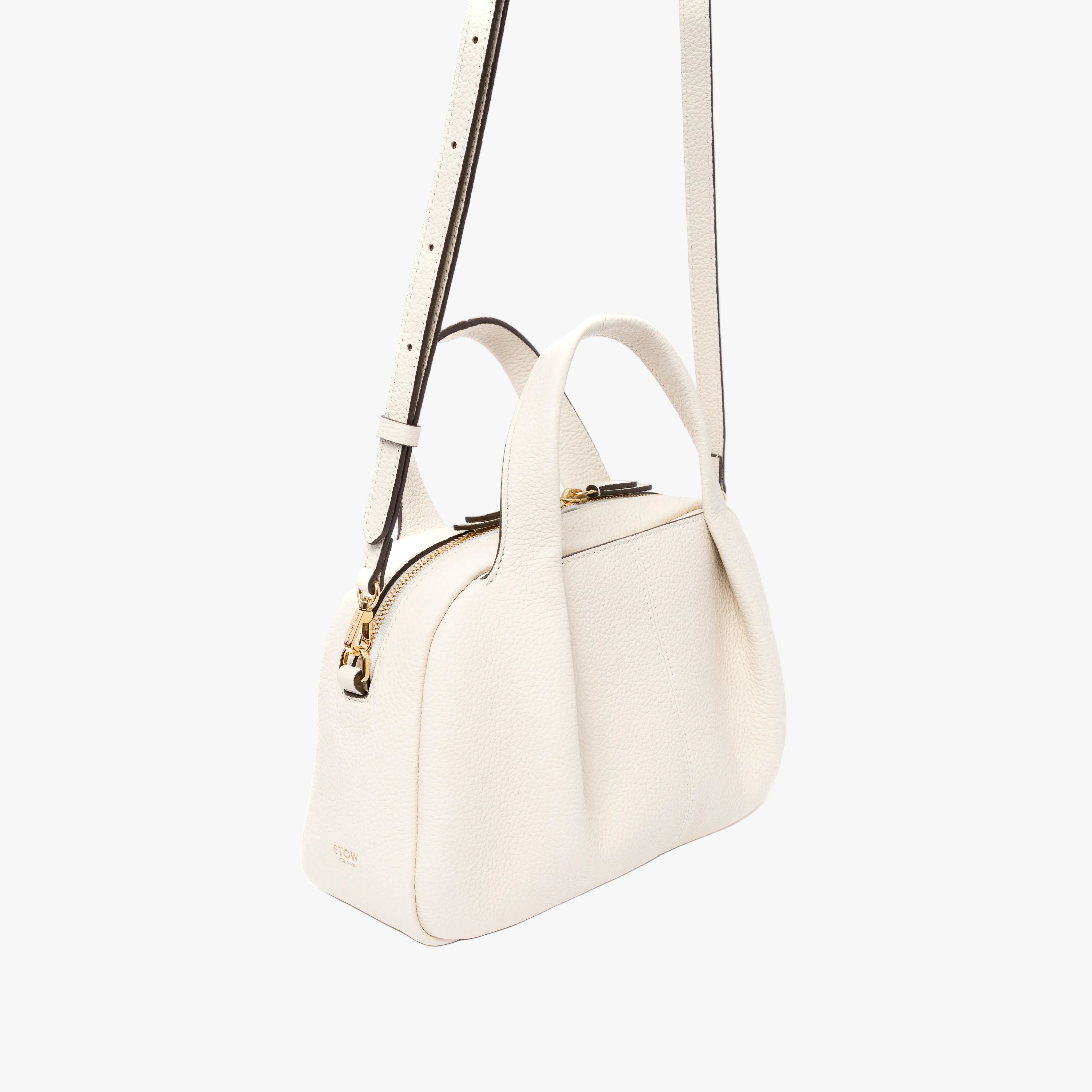 Off-axis view of STOW London's Mini Bowling bag in Spring Moon pebbled leather featuring a pleated front panel and gold-coated stainless steel hardware on a light grey background
