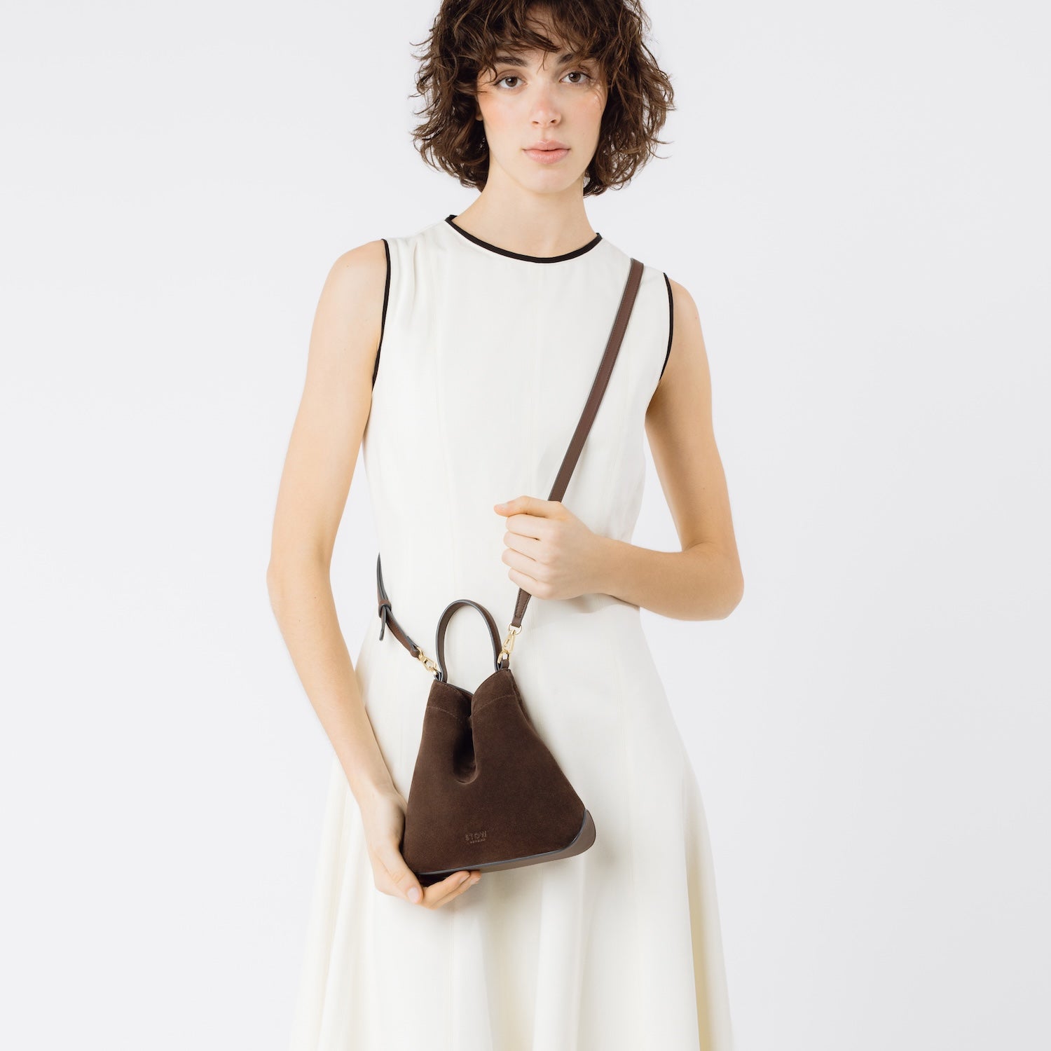 Model holding STOW London's Mini Curve in Chocolate suede leather crossbody by the detachable full leather strap