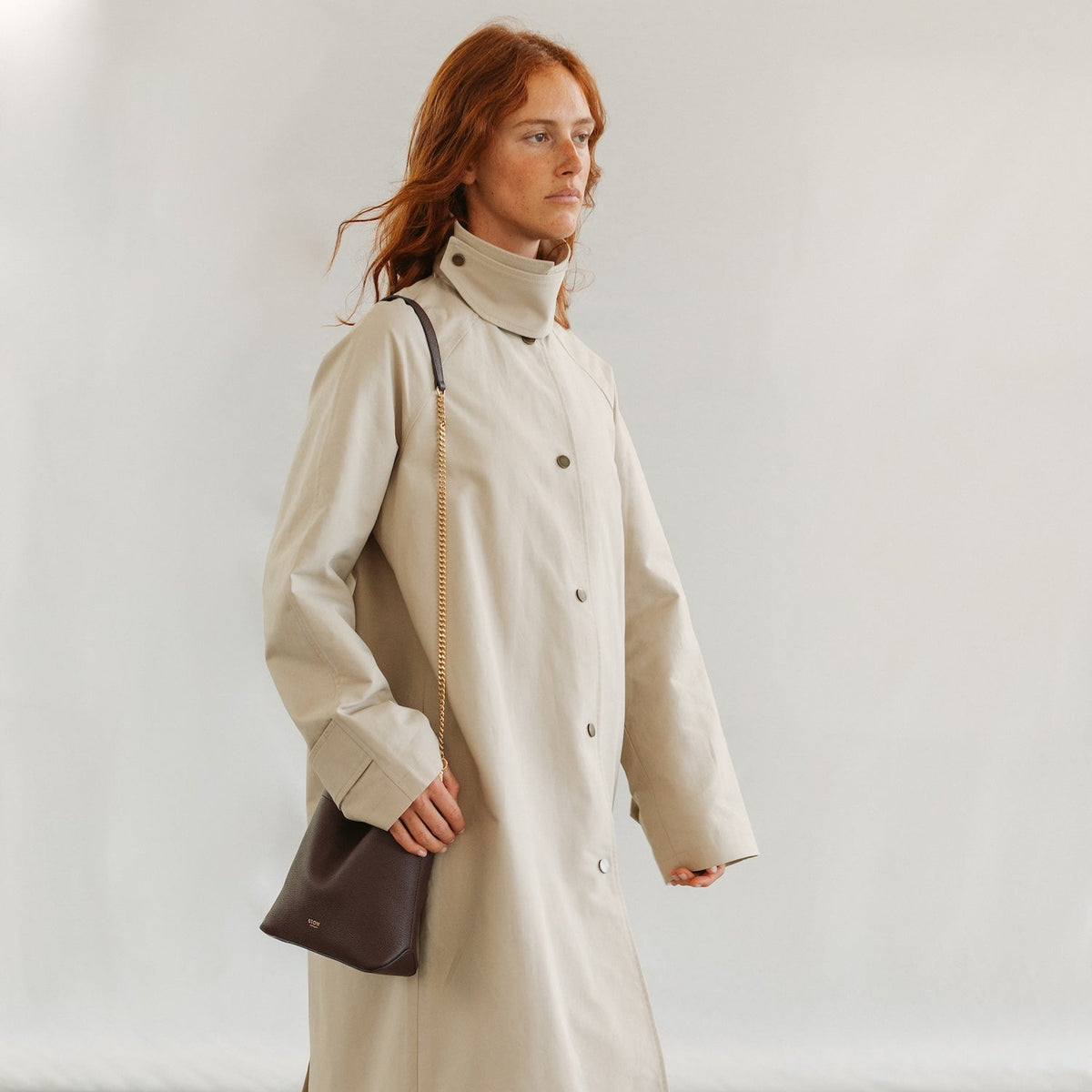 Model in a beige coat wearing STOW London's Mini Curve in Espresso pebbled leather crossbody featuring a gold plated chain strap