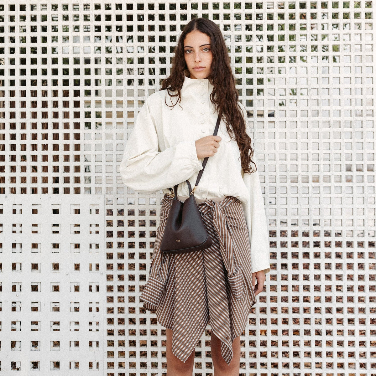 Model wearing STOW London's Mini Curve in Espresso pebbled leather crossbody featuring the detachable leather strap and gold-coated stainless steel hardware 