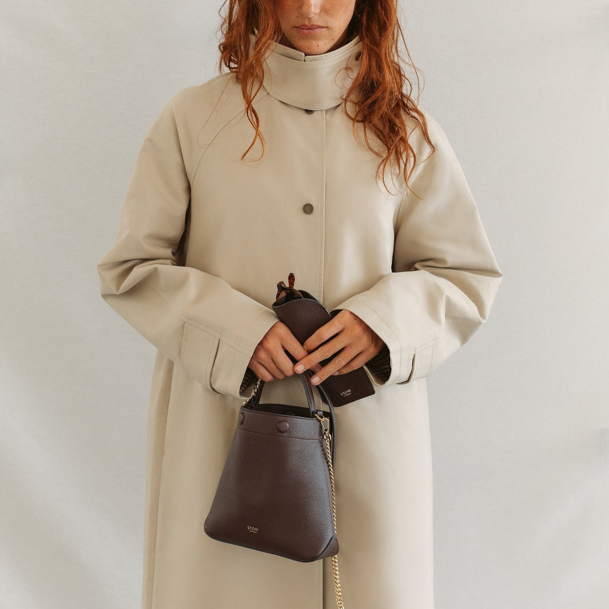 Model holding STOW London's Mini Curve in Espresso pebbled leather on her shoulder featuring the detachable gold chain strap and matching glasses case