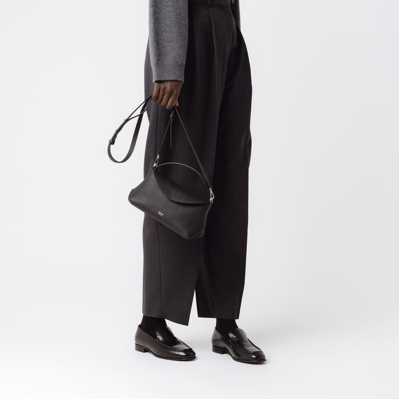Model holding STOW London's Mini East West in Black pebbled leather by its crossbody strap featuring silver stainless steel hardware on a white background