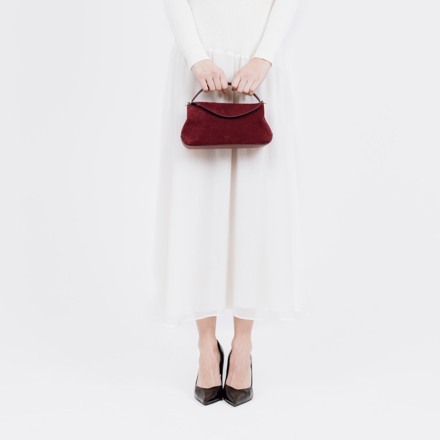 Model holding STOW London's Mini East West bag in Burgundy Suede leather by the top handle at her front featuring gold-coated stainless steel hardware on a white background