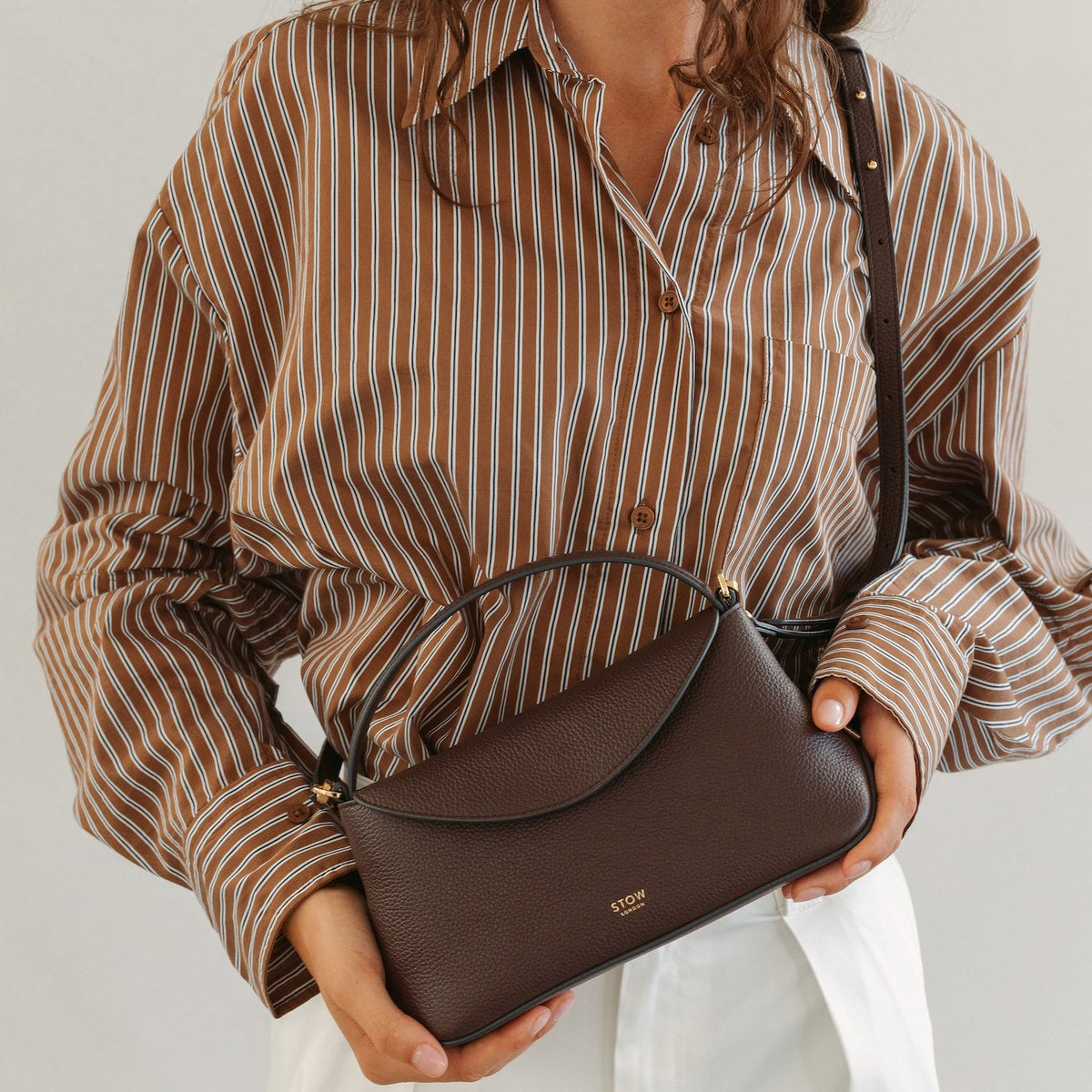 Model wearing STOW London's Mini East West in Espresso pebbled leather featuring the detachable crossbody strap and gold-coated stainless steel hardware 