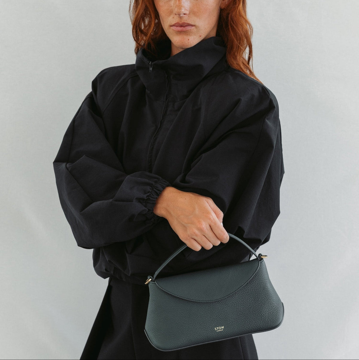 Model in a full black outfit holding STOW London's Mini East West in New Forest pebbled leather featuring gold-coated stainless steel hardware 