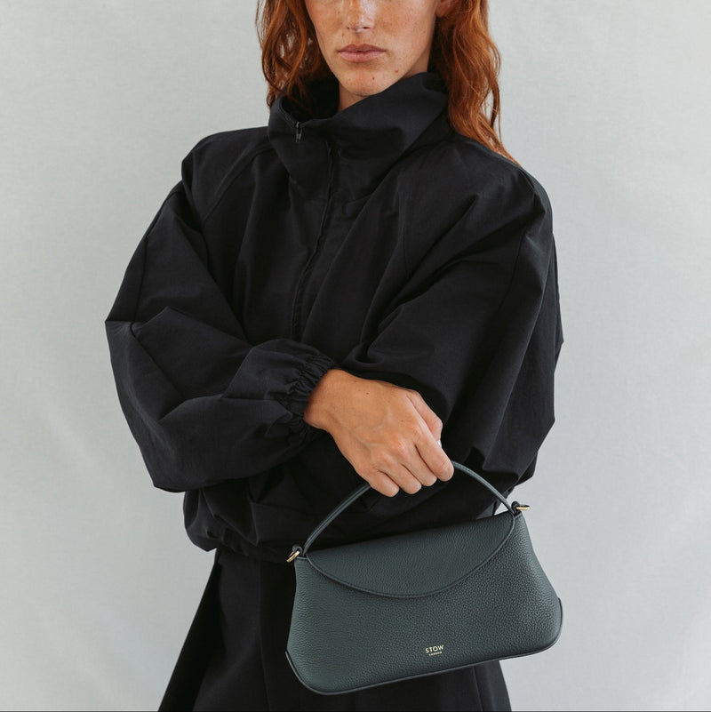 Model in a full black outfit holding STOW London's Mini East West in New Forest pebbled leather featuring gold-coated stainless steel hardware 