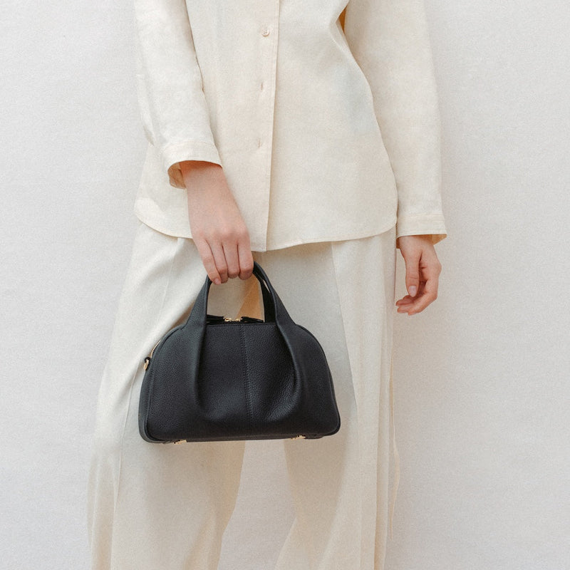 Model holding STOW London's Mini Bowling in Black pebbled leather featuring a pleated front panel and gold-coated stainless steel hardware