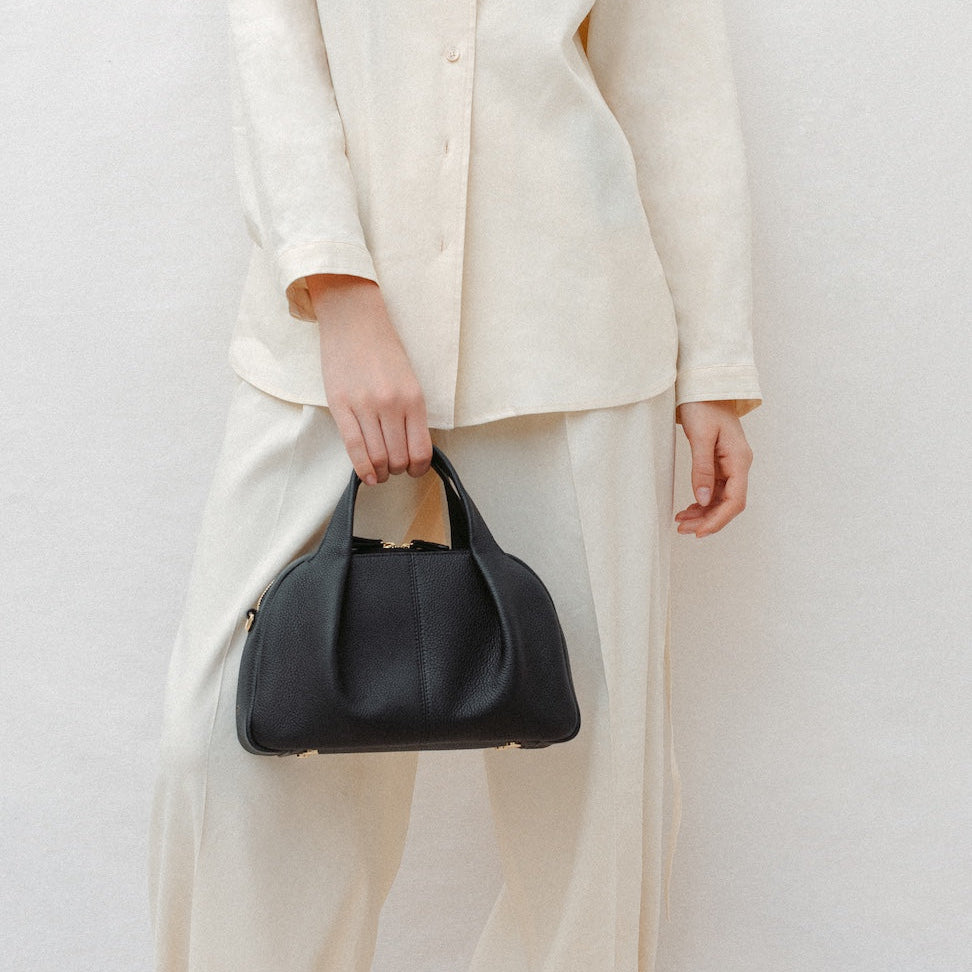 Model holding STOW London's Mini Bowling in Black pebbled leather featuring a pleated front panel and gold-coated stainless steel hardware