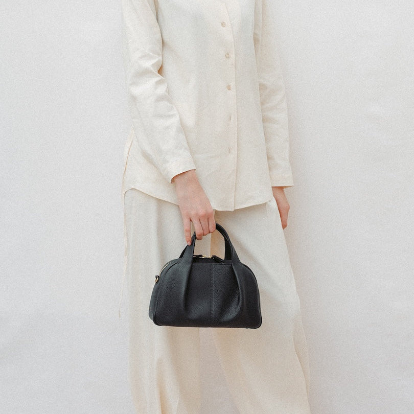 Model holding STOW London's Mini Bowling in Black pebbled leather featuring a pleated front panel and gold-coated stainless steel hardware