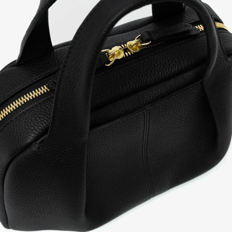 Close up of STOW London's Mini Bowling bag in Black pebbled leather featuring a pleated front panel with the external slip pocket in-between, zip closure, STOW London embossed pull tabs and gold-coated stainless steel hardware on a light grey background