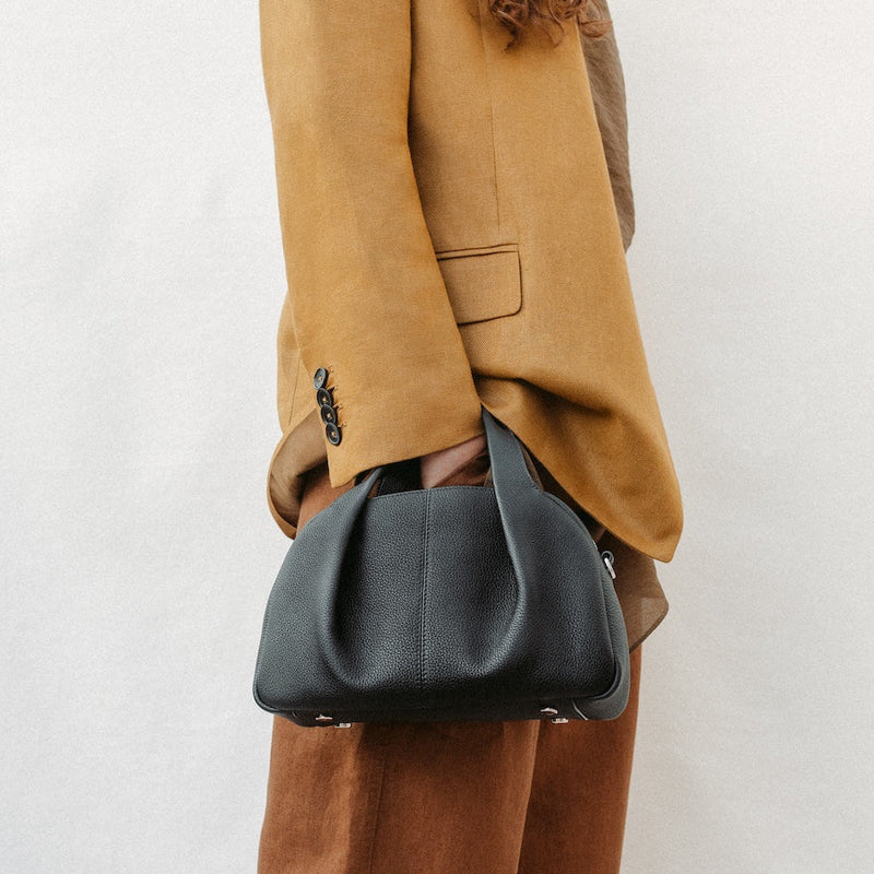 Model holding STOW London's Mini Bowling in Black pebbled leather on her wrist featuring a pleated front panel and silver stainless steel hardware