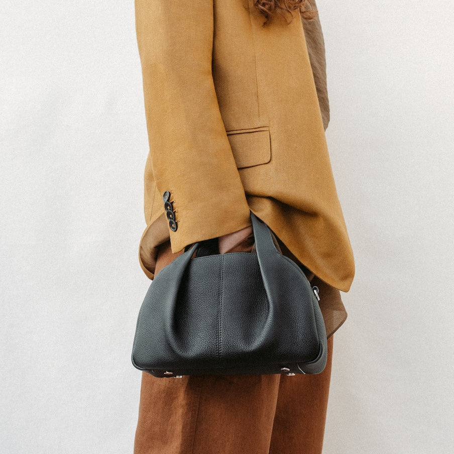 Model holding STOW London's Mini Bowling in Black pebbled leather on her wrist featuring a pleated front panel and silver stainless steel hardware
