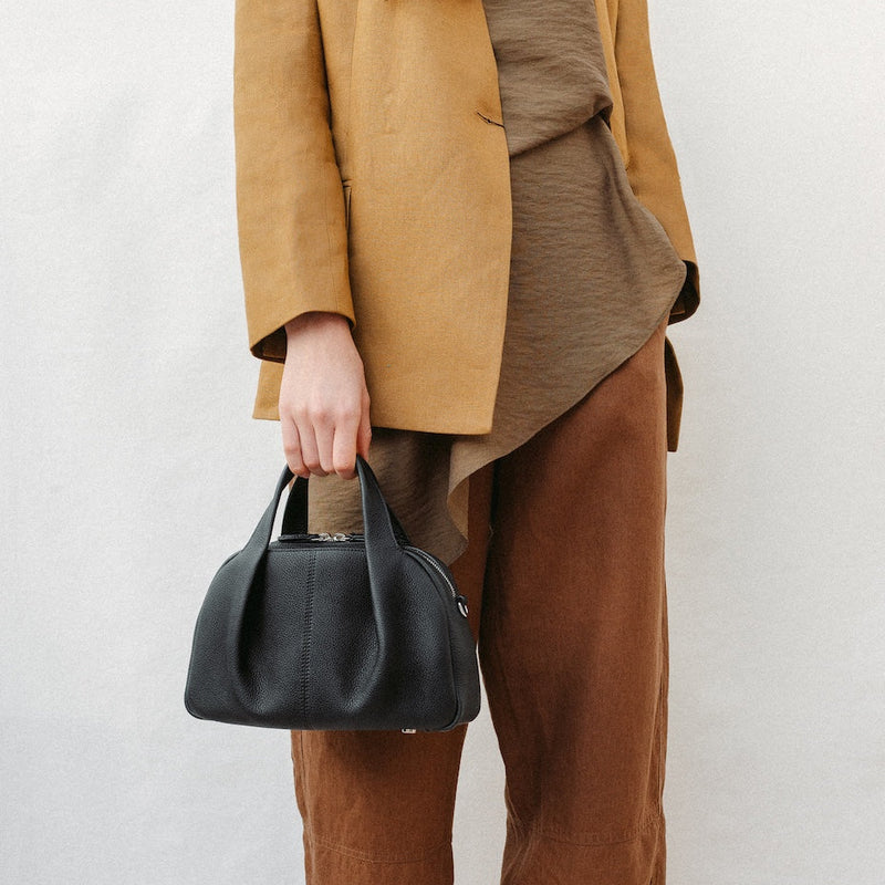 Model holding STOW London's Mini Bowling in Black pebbled leather at her side featuring a pleated front panel and silver stainless steel hardware