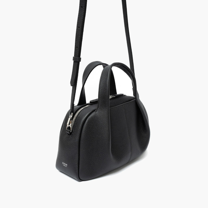 Off-axis view of STOW London's Mini Bowling bag in Black pebbled leather featuring a pleated front panel, detachable crossbody strap and silver stainless steel hardware on a light grey background