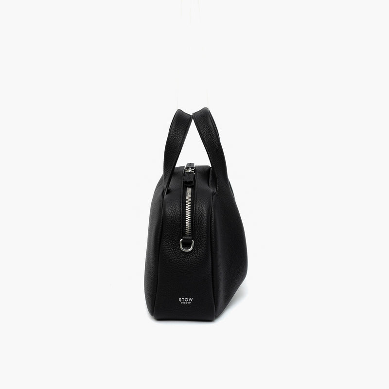 Side view of STOW London's Mini Bowling bag in Black pebbled leather featuring a silver stainless steel hardware, zip and small logo on a light grey background