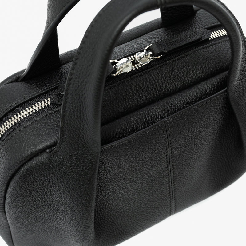 Close up of STOW London's Mini Bowling bag in Black pebbled leather featuring a pleated front panel with the external slip pocket in-between, zip closure, STOW London embossed pull tabs and silver stainless steel hardware on a light grey background