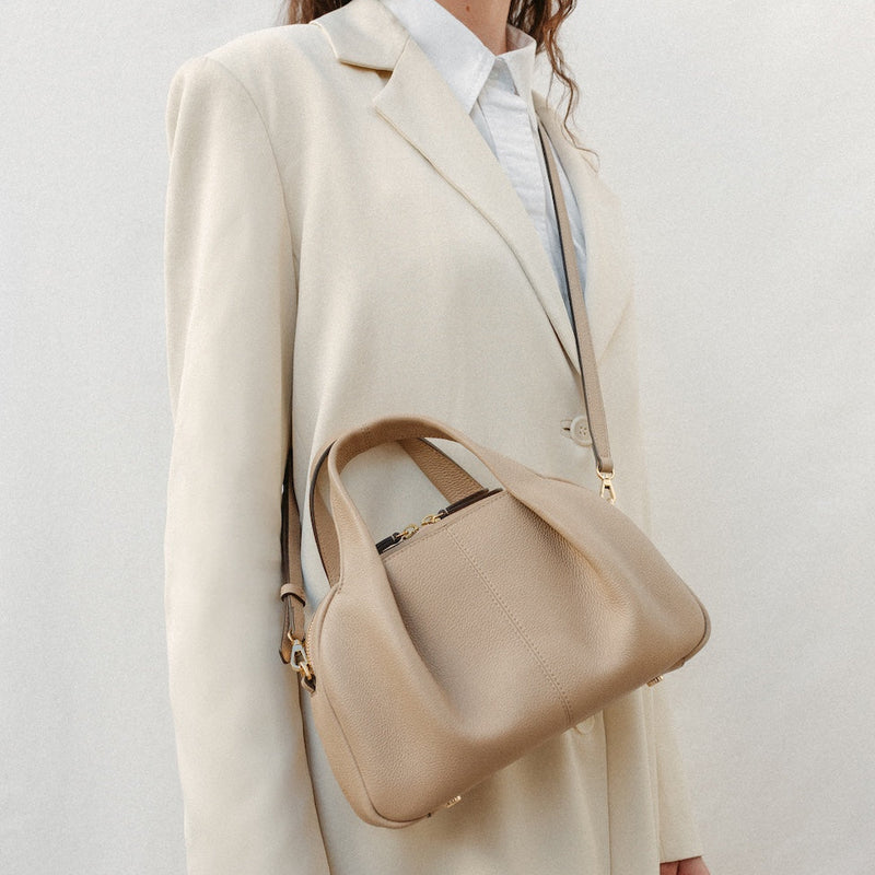 Model wearing STOW London's Mini Bowling bag in Camel pebbled leather crossbody featuring a pleated front panel, detachable crossbody strap and gold-coated stainless steel hardware