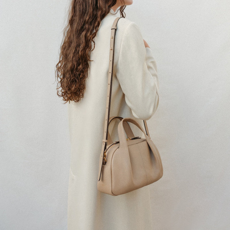 Model wearing STOW London's Mini Bowling bag in Camel pebbled leather over her shoulder featuring a pleated front panel, detachable crossbody strap and gold-coated stainless steel hardware 