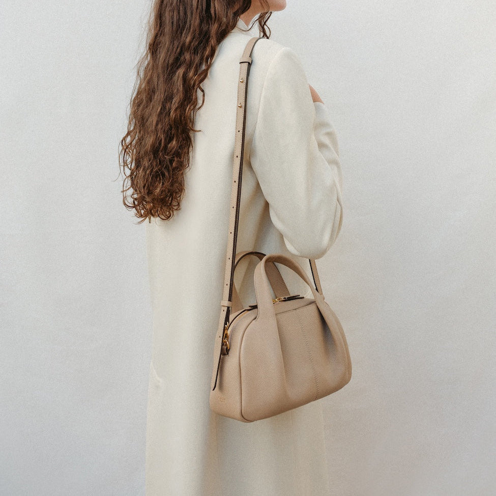 Model wearing STOW London's Mini Bowling bag in Camel pebbled leather over her shoulder featuring a pleated front panel, detachable crossbody strap and gold-coated stainless steel hardware 