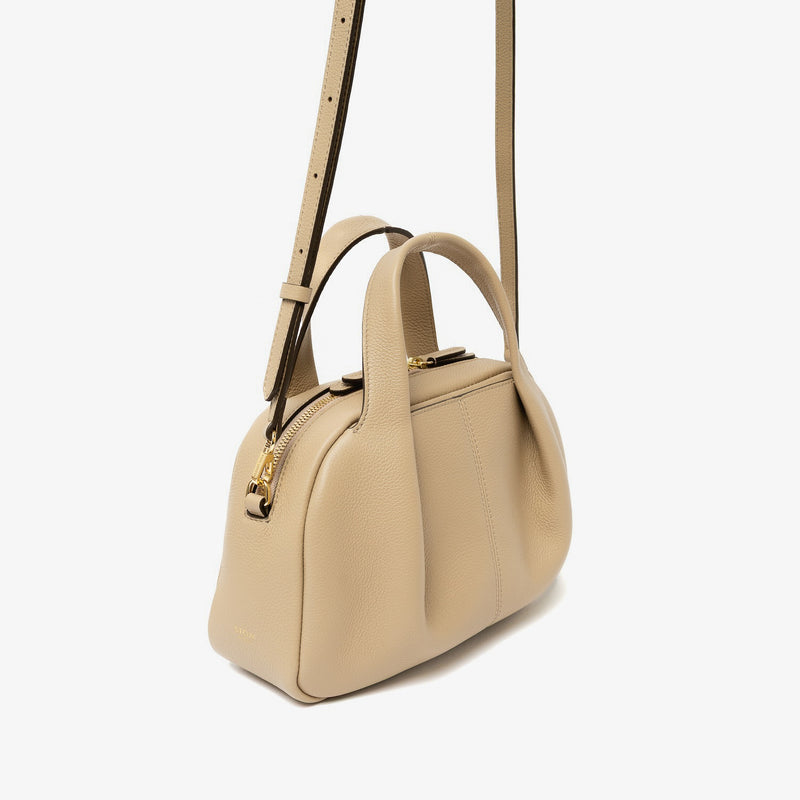 Off-axis view of STOW London's Mini Bowling bag in Camel pebbled leather featuring a pleated front panel, detachable crossbody strap and gold-coated stainless steel hardware on a light grey background