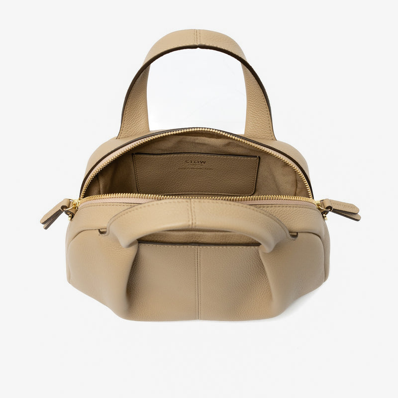 STOW London's Mini Bowling bag in Camel pebbled leather with zip open showing the light beige cotton interior and slip pocket featuring gold-coated stainless steel hardware on a light grey background