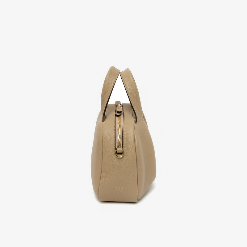 Side view of STOW London's Mini Bowling bag in Camel pebbled leather featuring the zip closure and small gold logo on a light grey background