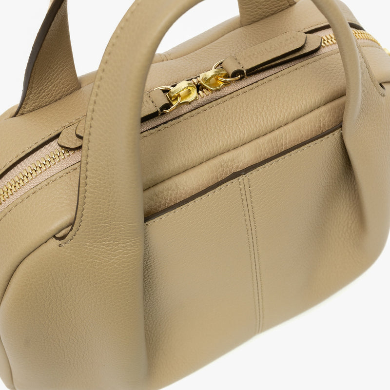 Close up of STOW London's Mini Bowling bag in Camel pebbled leather featuring a pleated front panel, external slip pocket and gold-coated stainless steel hardware on the zip closure on a light grey background