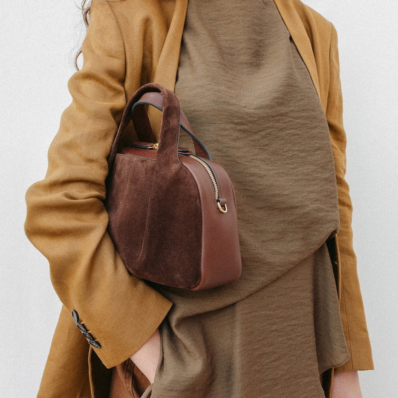 Model holding STOW London's Mini Bowling in Chocolate suede leather featuring a pleated front panel and gold-coated stainless steel hardware