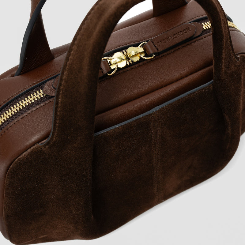 Close up of STOW London's Mini Bowling bag in Chocolate suede and smooth leather featuring a pleated front panel with slip pocket in-between, zip closure and STOW London embossed pull tabs on a light grey background