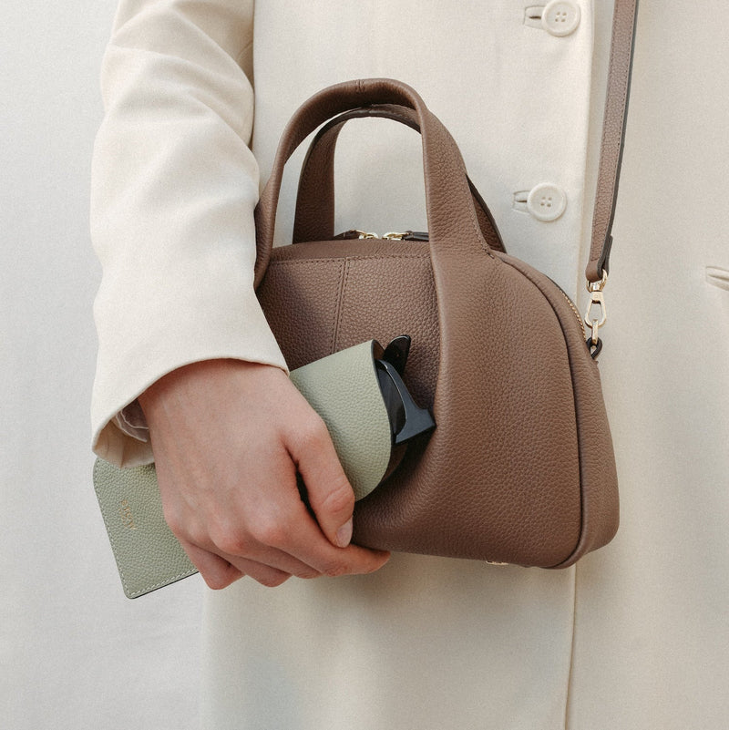 Model holding STOW London's Glasses Case in Pale Green pebbled leather and the Mocha Mini Bowling bag featuring a gold logo 