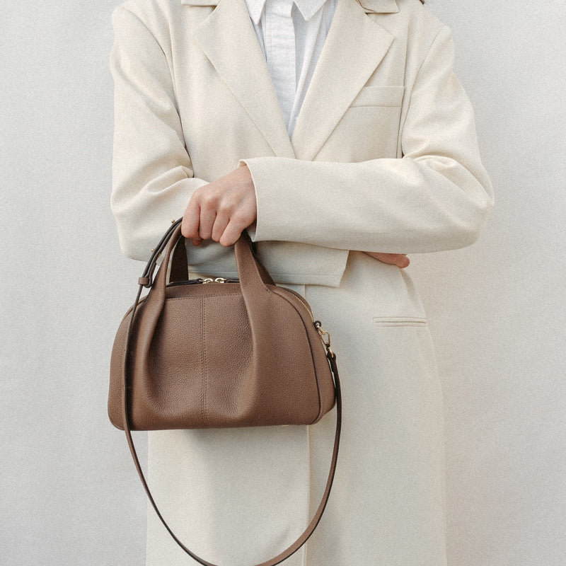 Model holding STOW London's Mini Bowling bag in Mocha pebbled leather featuring the detachable crossbody strap, pleated front panel and gold-coated stainless steel hardware
