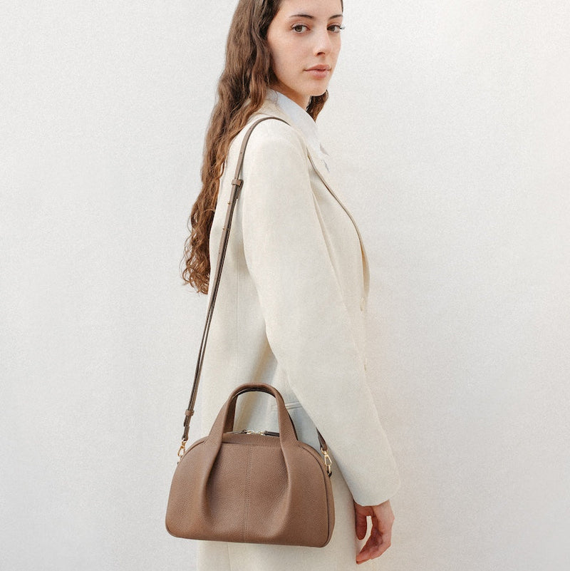 Model wearing STOW London's Mini Bowling bag in Mocha pebbled leather over her shoulder featuring a pleated front panel, detachable crossbody strap and gold-coated stainless steel hardware