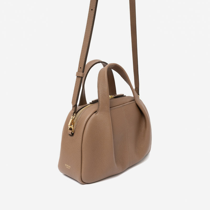 Off-axis view of STOW London's Mini Bowling bag in Mocha pebbled leather featuring a pleated front panel, the detachable crossbody strap and gold-coated stainless steel hardware on a light grey background