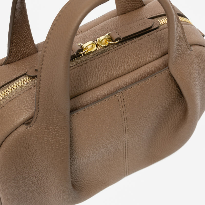 Close up of STOW London's Mini Bowling bag in Mocha pebbled leather featuring the pleated front panel with external pocket in-between, gold-coated stainless steel hardware, zip closure and STOW London embossed pull tabs on a light grey background
