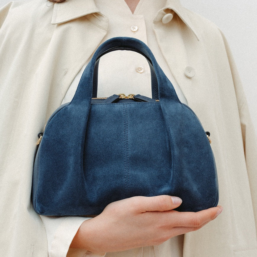 Model holding STOW London's Mini Bowling in Navy suede leather featuring a pleated front panel and gold-coated stainless steel hardware