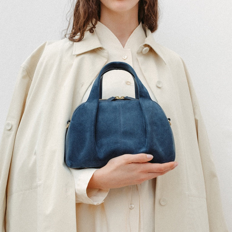 Model holding STOW London's Mini Bowling in Navy suede leather featuring a pleated front panel and gold-coated stainless steel hardware