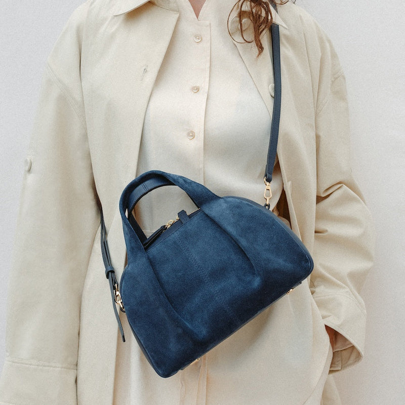 Model wearing STOW London's Mini Bowling in Navy suede leather crossbody featuring a pleated front panel and gold-coated stainless steel hardware