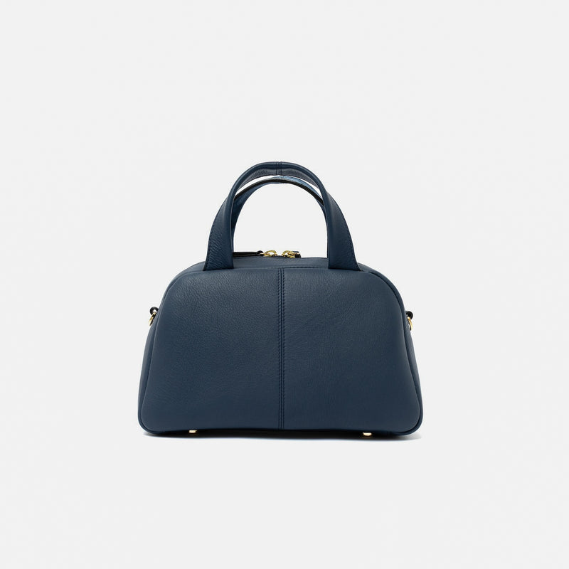 Shot of the smooth leather back panel of STOW London's Mini Bowling bag in Navy featuring gold-coated stainless steel hardware and four feet on the base on a light grey background