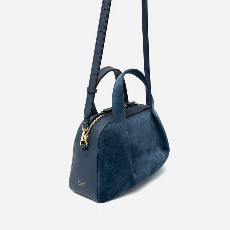 Off-axis view of STOW London's Mini Bowling bag in Navy suede and smooth leather featuring its detachable crossbody strap, a pleated front panel and gold-coated stainless steel hardware on a light grey background