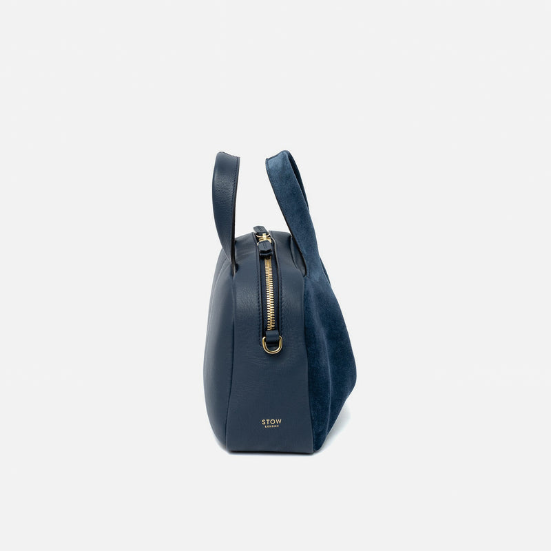 Side view of STOW London's Mini Bowling bag in Navy suede and smooth leather featuring gold-coated stainless steel hardware and small STOW logo on a light grey background
