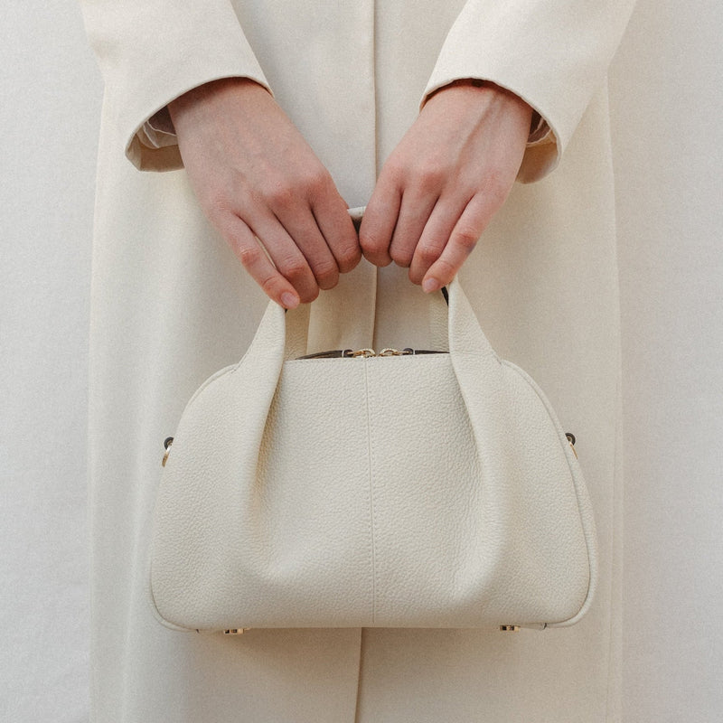 Model holding STOW London's Mini Bowling bag in Spring Moon pebbled leather by top handle featuring a pleated front panel and gold-coated stainless steel hardware 