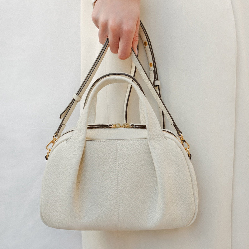 Close up of model holding STOW London's Mini Bowling bag in Spring Moon pebbled leather by its detachable crossbody strap at her side featuring a pleated front panel and gold-coated stainless steel hardware 
