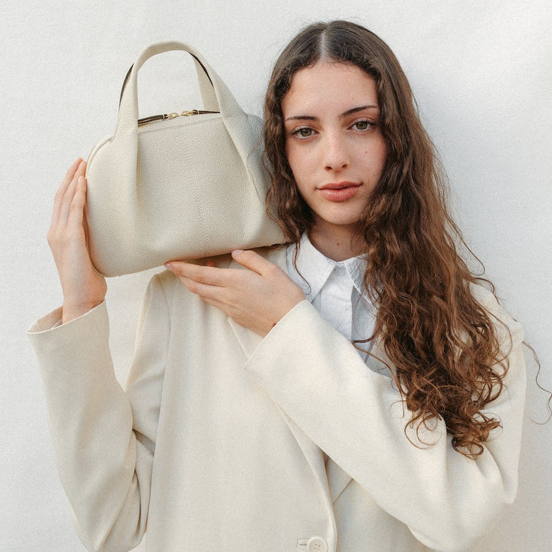 Model holding STOW London's Mini Bowling bag in Spring Moon pebbled leather featuring a pleated front panel and gold-coated stainless steel hardware