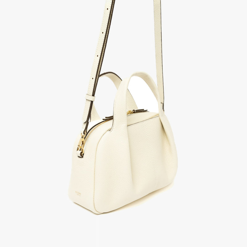 Off-axis view of STOW London's Mini Bowling bag in Spring Moon pebbled leather featuring a pleated front panel, detachable crossbody strap and gold-coated stainless steel hardware on a light grey background